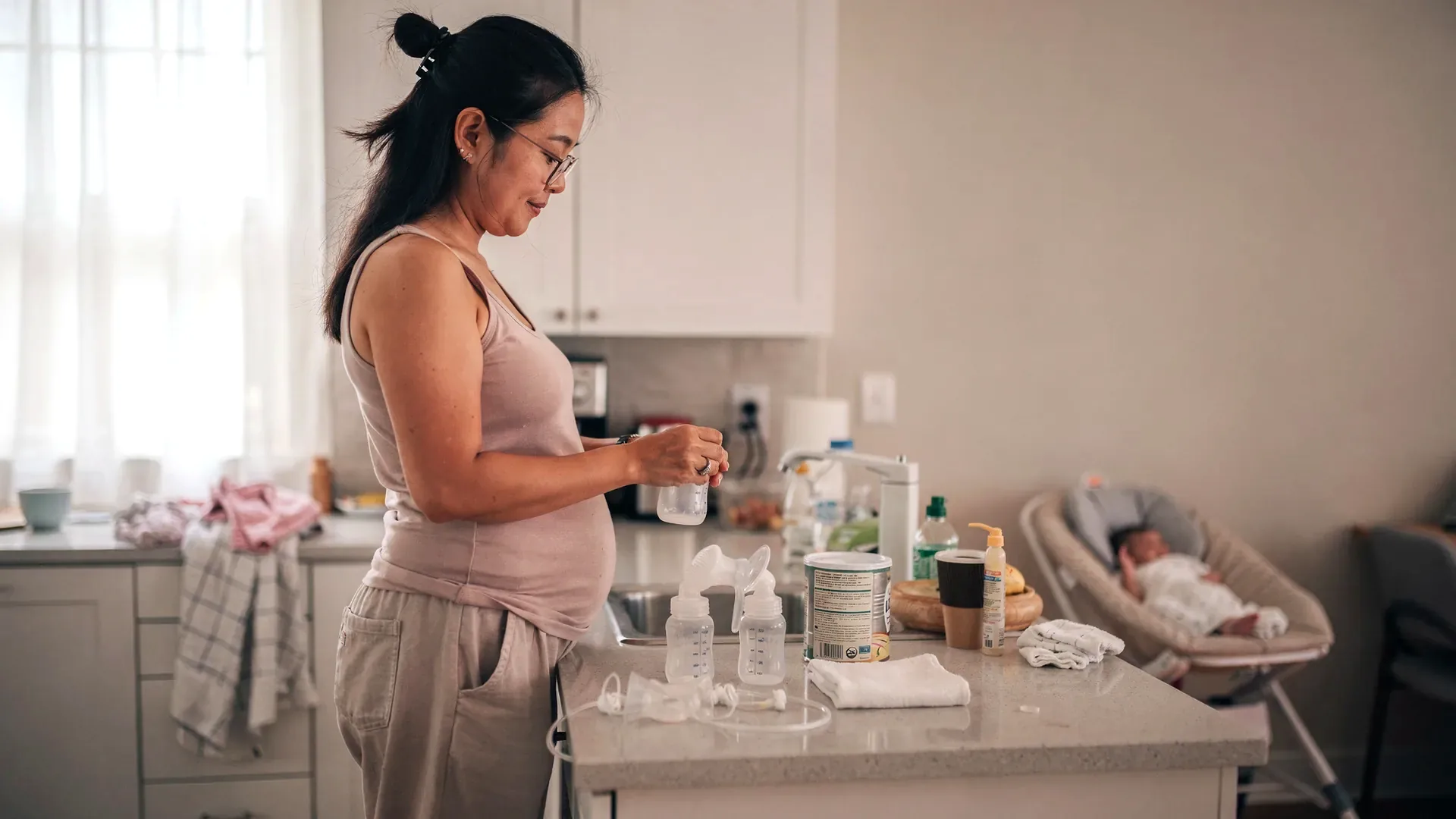 An East Asian postpartum woman with long black hair in a claw clip and tall lean-soft body prepares a bottle at a kitchen counter with pump parts and formula nearby.