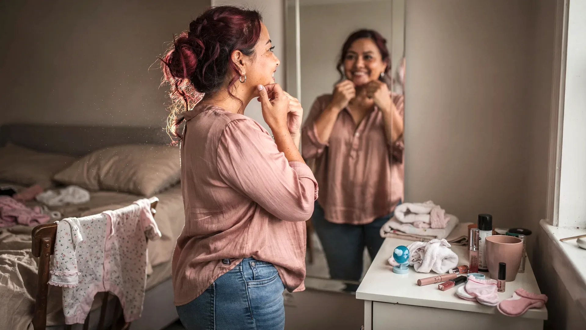 A mixed Indigenous and white postpartum woman with dyed burgundy curls and petite soft body clasps earrings in a bedroom mirror with baby clothes and makeup nearby.