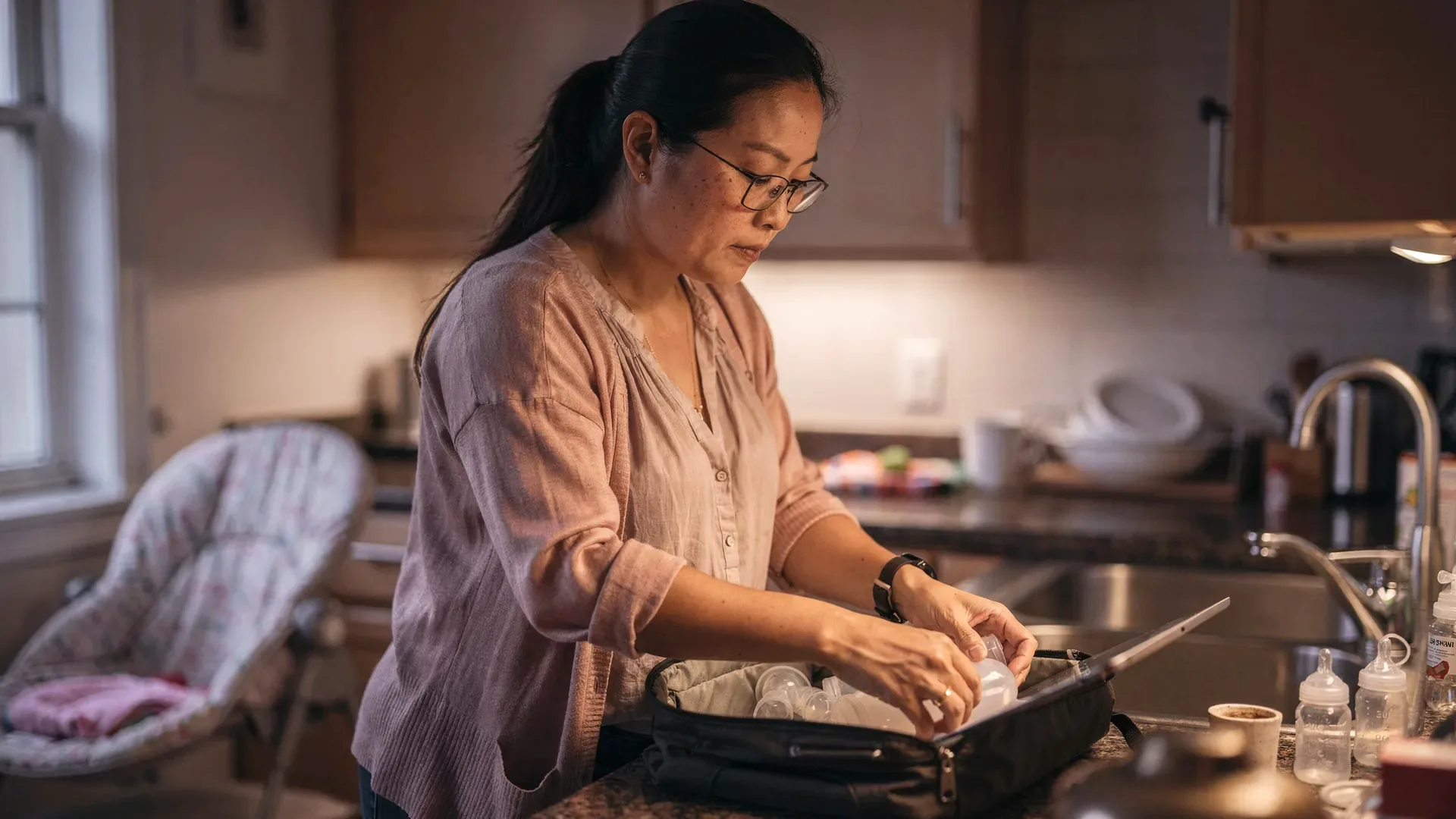 A mixed East Asian and white postpartum woman with long dark hair in a low ponytail and tall average body packs a work bag with pump parts and a laptop in dim morning light.