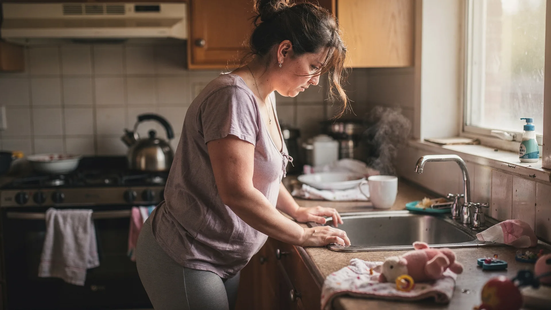 A woman grips a kitchen counter beside scattered toys and a kettle after a hard moment.