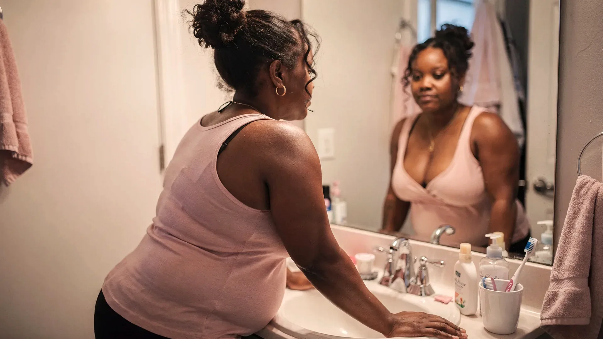 A Black postpartum woman with curly natural hair and athletic-soft build leans over a bathroom sink and looks steadily into the mirror in warm morning light.