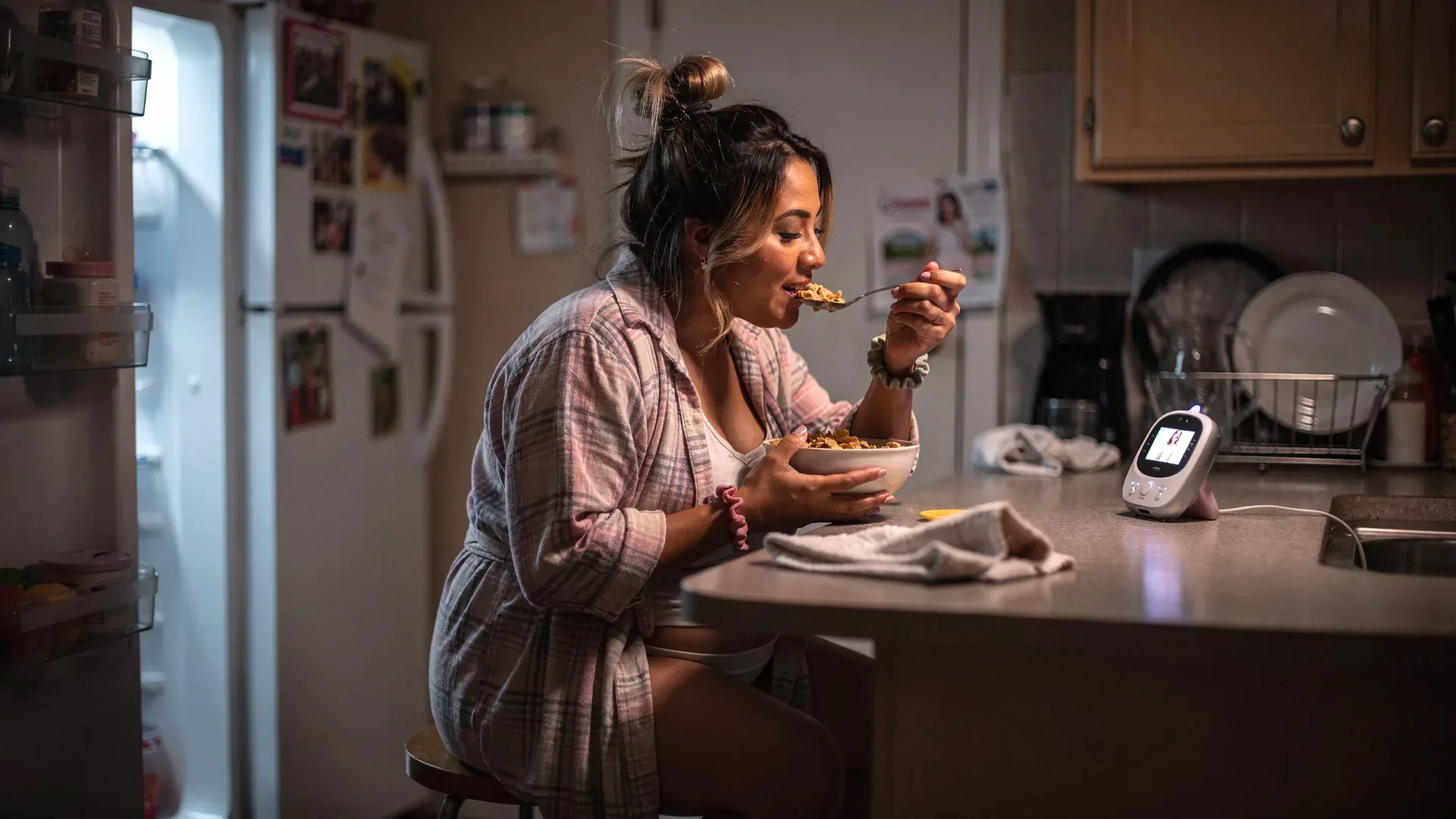 A Latina postpartum woman with messy bun with blonde streak and average postpartum body in a robe eats cereal at night in a kitchen lit by the open fridge.