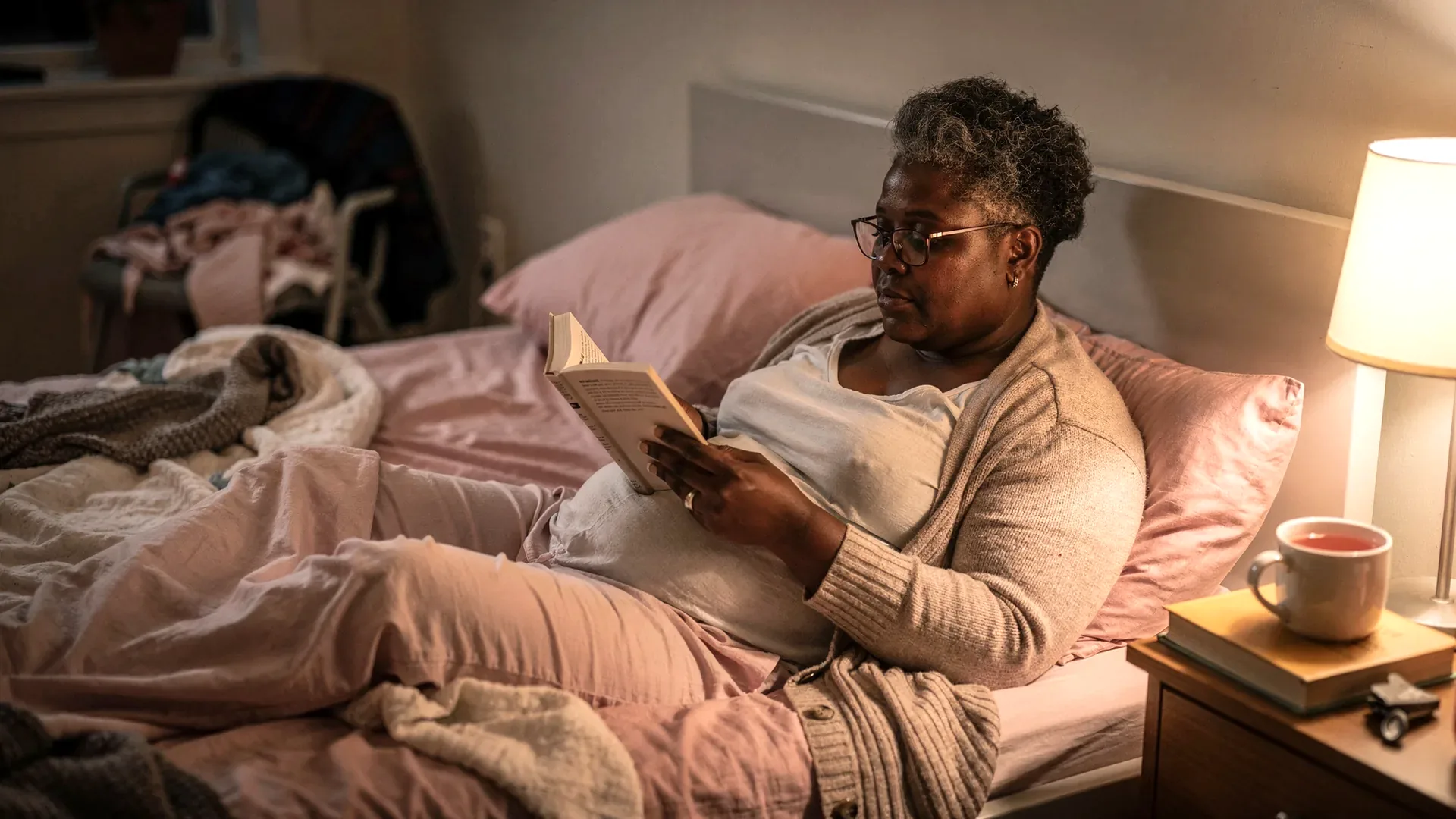 A Black postpartum woman with short gray natural curls and average body reads alone in a dim bedroom while the other side of the bed is softly out of focus.