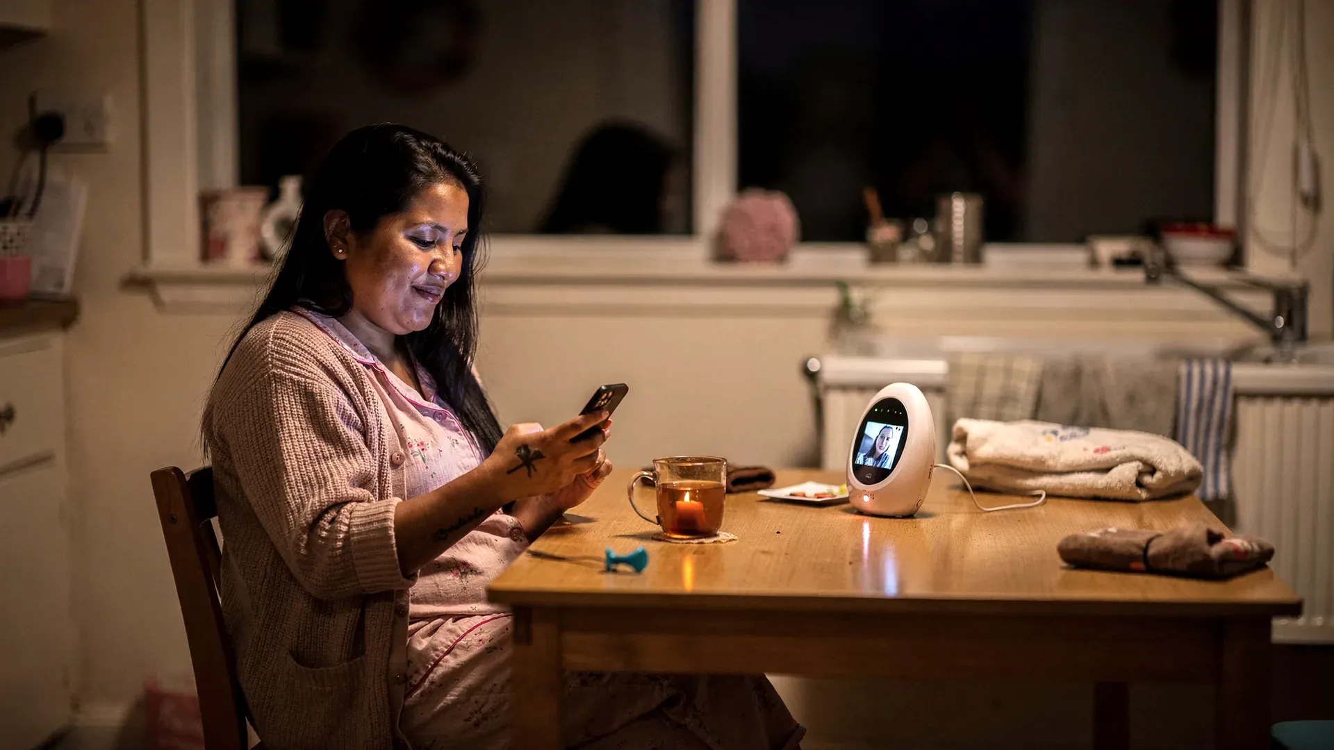 An Indigenous postpartum woman with long dark straight hair and average soft body sits alone at a kitchen table at night looking at an old photo on her phone.