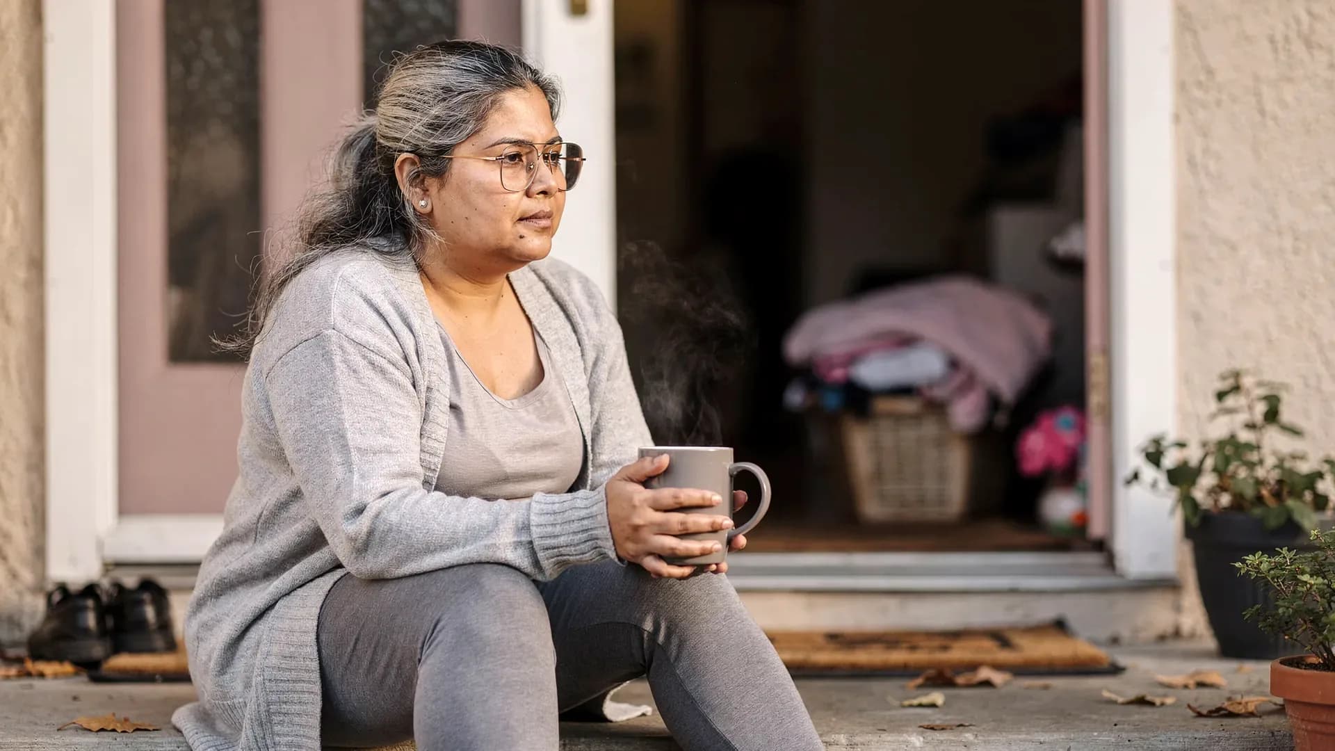A Latina postpartum woman with long silver-streaked hair and athletic-soft body sits alone on a front step with coffee while the door is cracked behind her.
