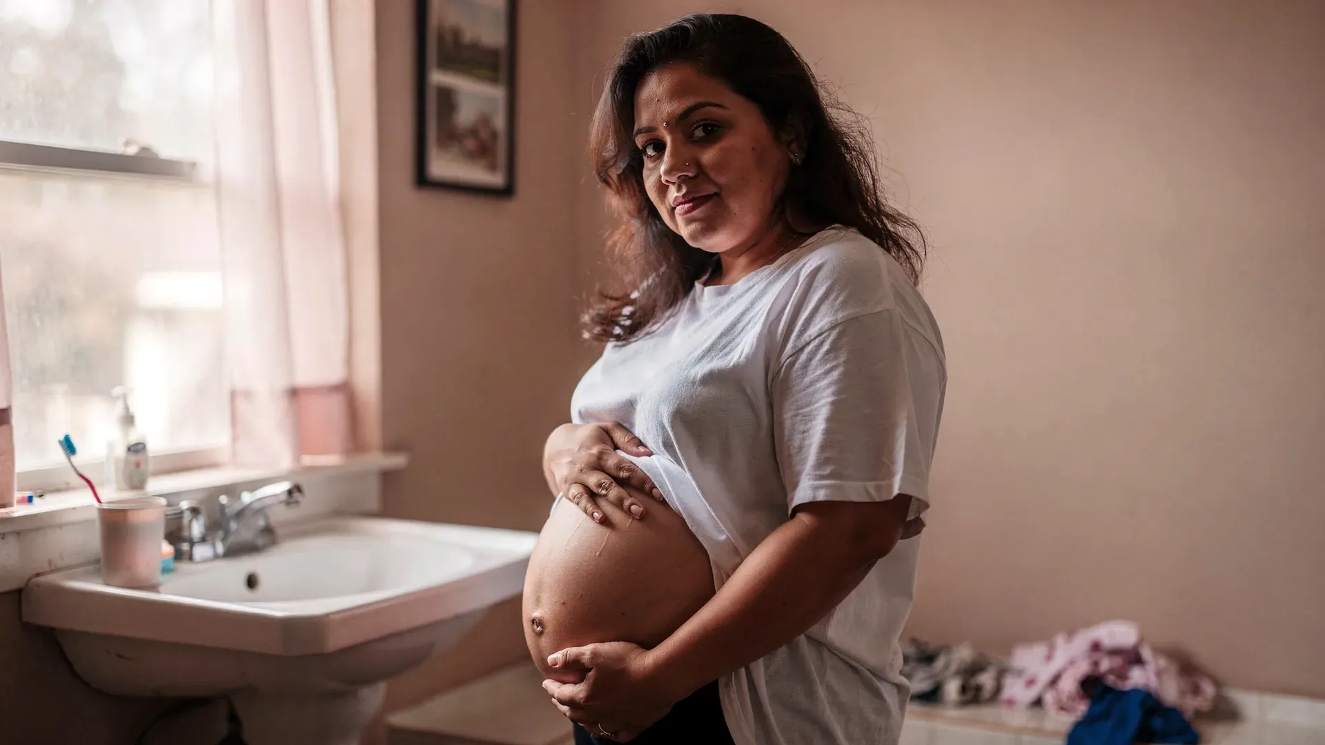 A Latina postpartum woman with long dark wavy hair and soft postpartum midsection in an oversized t-shirt stands by a bathroom mirror with one hand resting on her belly.