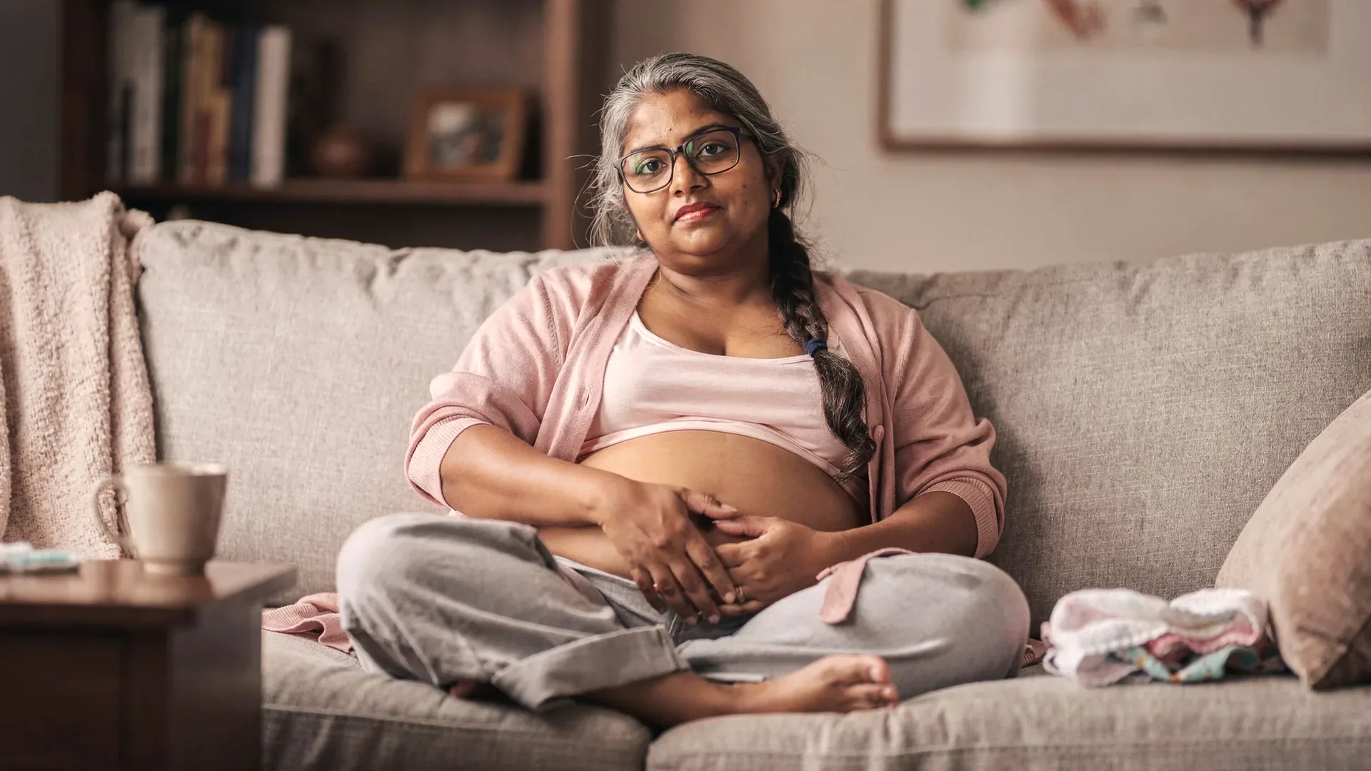 A South Asian postpartum woman with gray-streaked braid and petite soft body rests on a couch in a soft cardigan with laundry and a coffee mug nearby.
