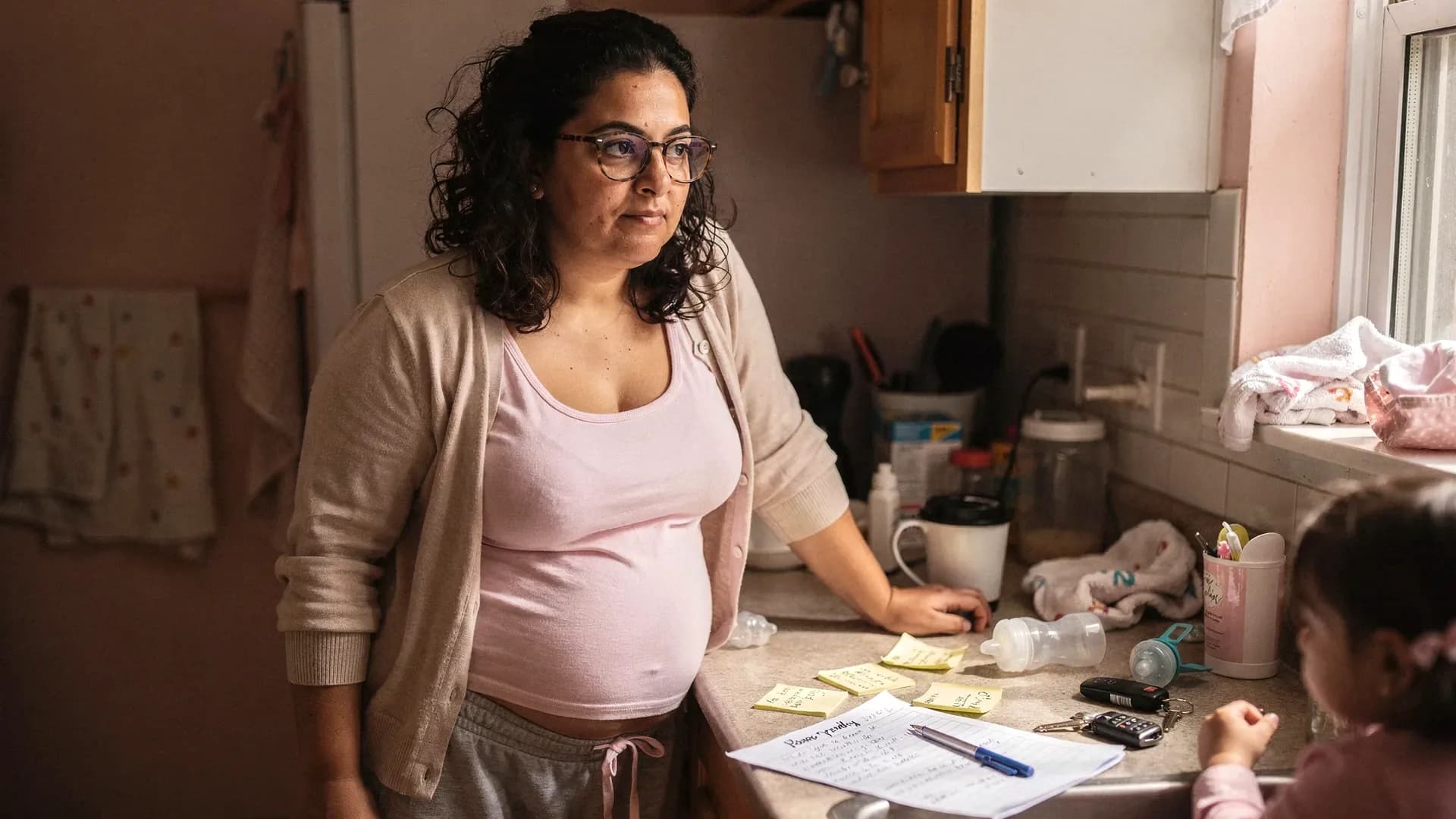 A tired Middle Eastern postpartum woman with curly dark shoulder-length hair and average curvy body stands at a kitchen counter covered with notes, keys, and bottle parts.