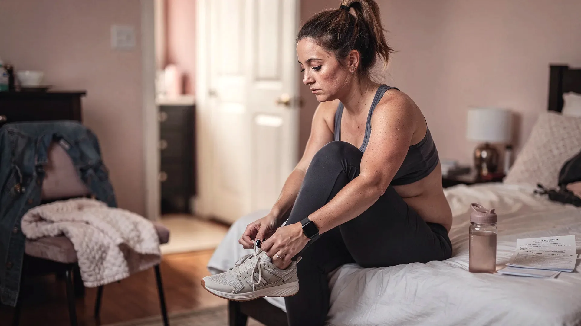 A white postpartum woman with high ponytail and athletic postpartum body sits on the edge of a bed tying her sneakers beside appointment notes.