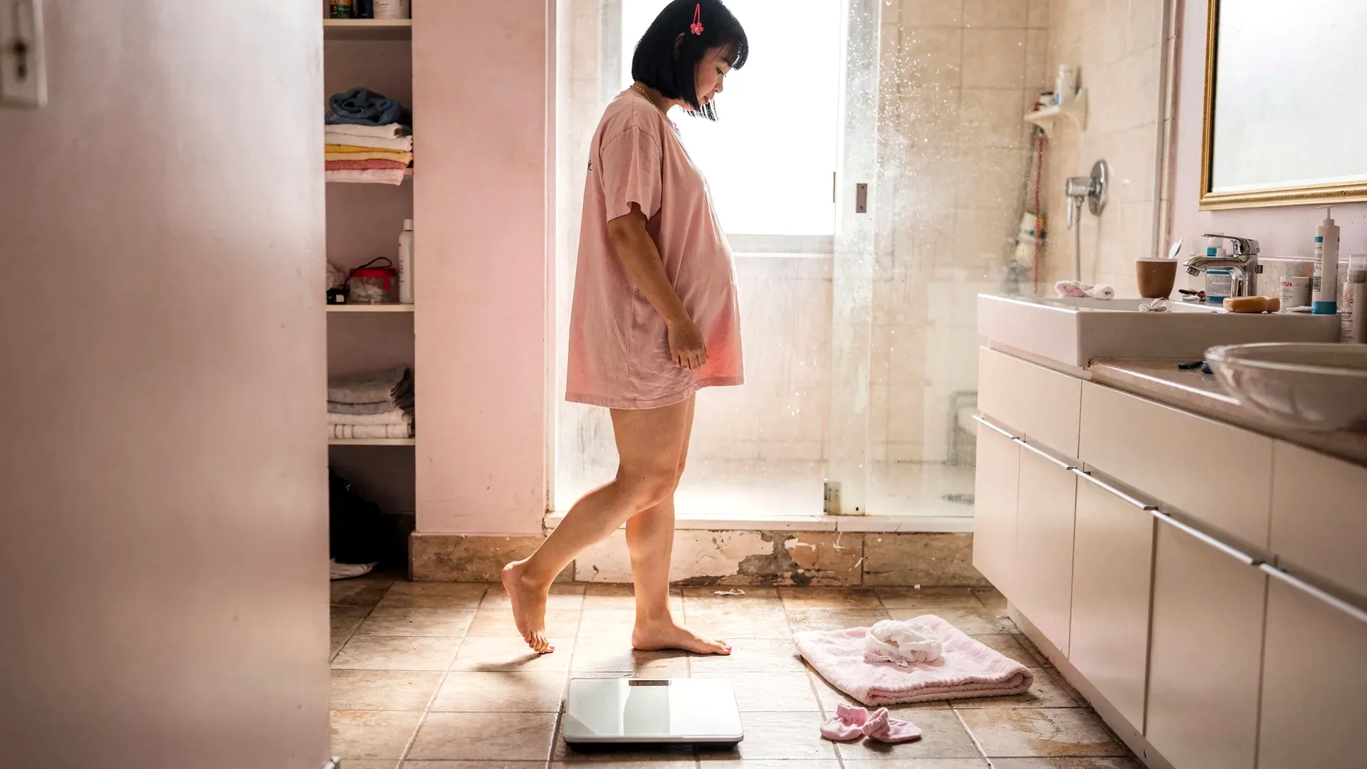 An East Asian postpartum woman with blunt black bob and petite body steps around a bathroom scale with a baby towel and small socks nearby.