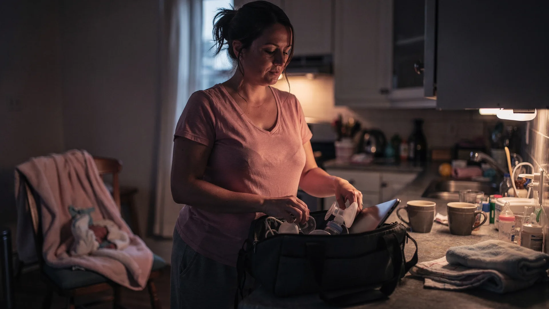 A woman packs a work bag with pump parts and a laptop in dim morning light.