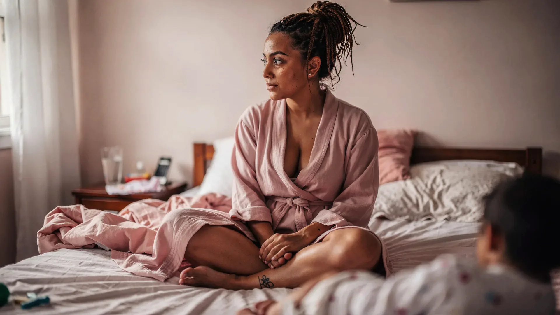 A Latina postpartum woman with loose braids and soft postpartum body sits thoughtfully on a bed in a robe under soft lamplight.