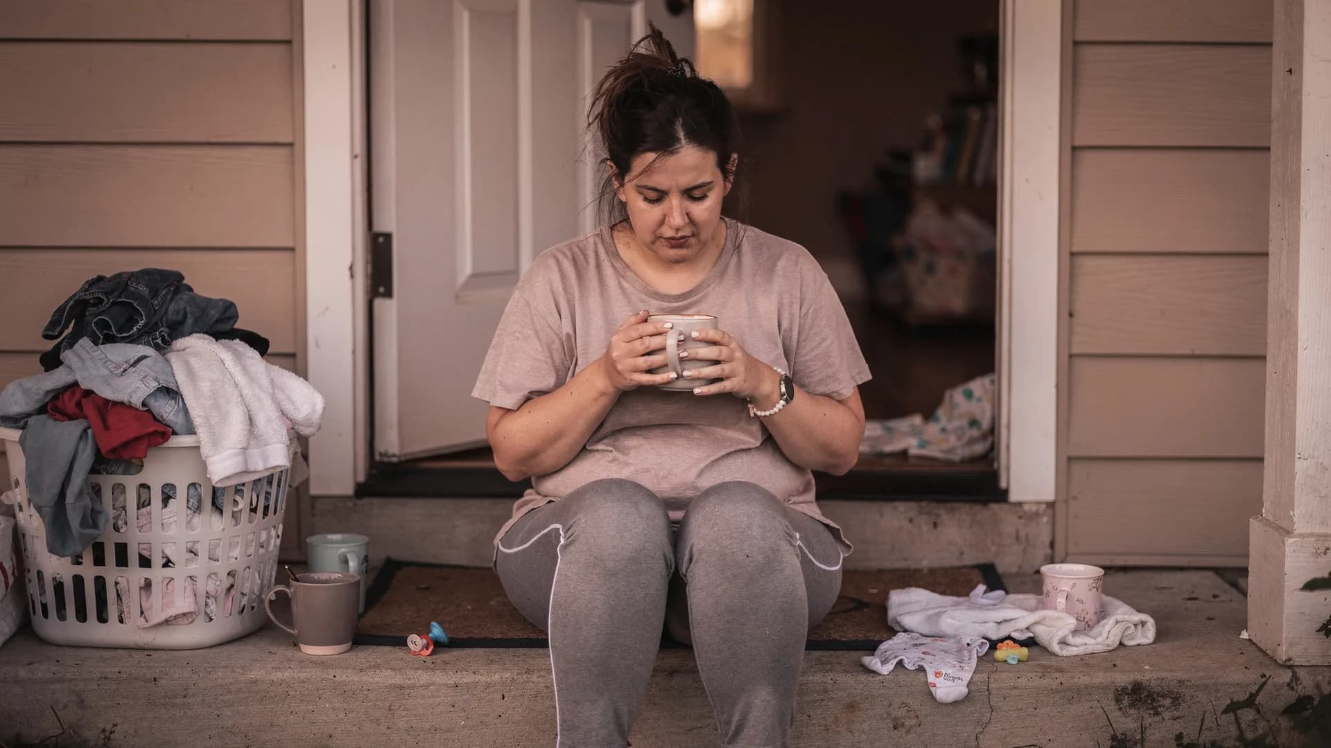 A woman sits alone on a front step with coffee while the door is cracked behind her.
