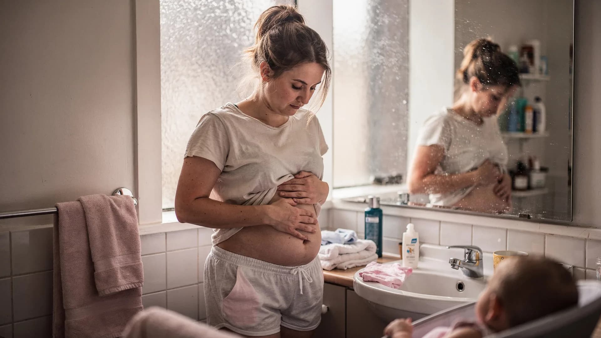 A woman gently lifts her shirt hem in a bathroom mirror while looking at her healing body.