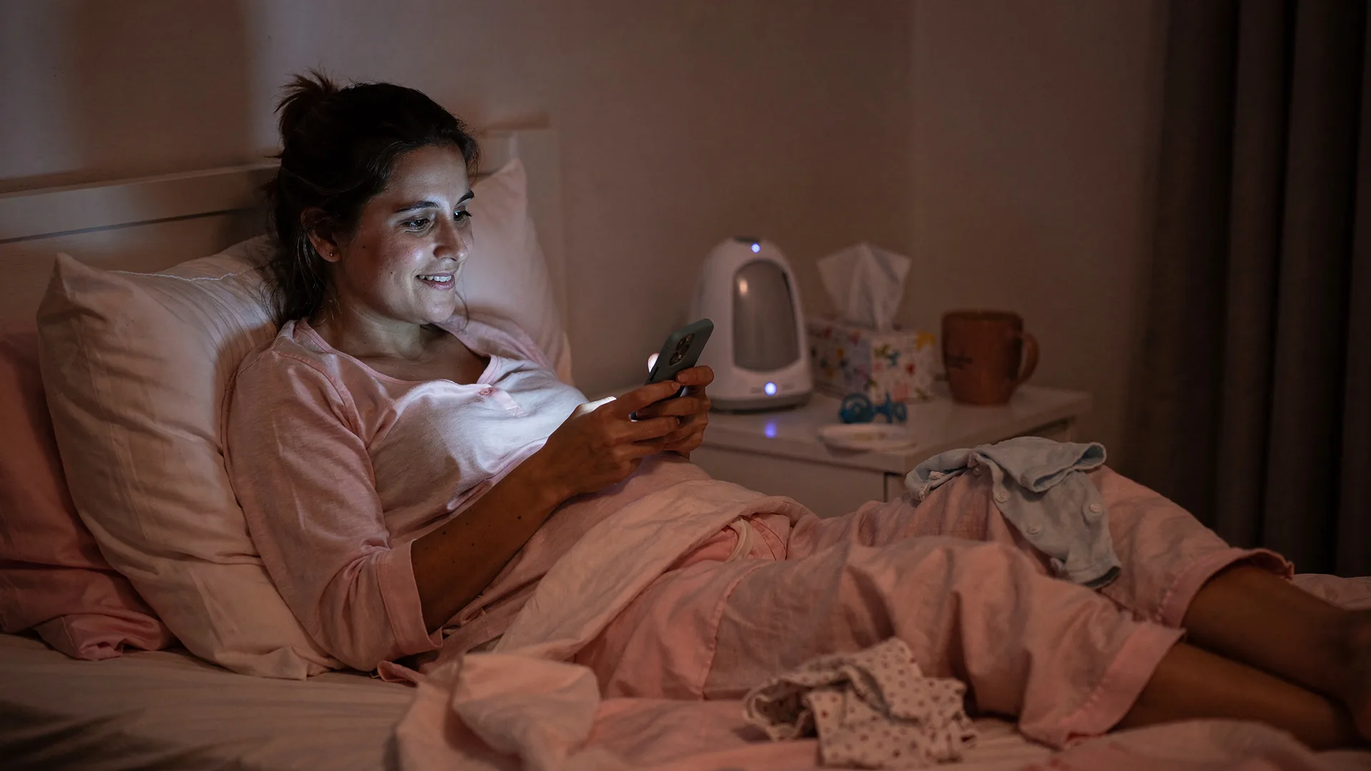 A tired woman smiles at her phone in bed at night with a baby monitor nearby.