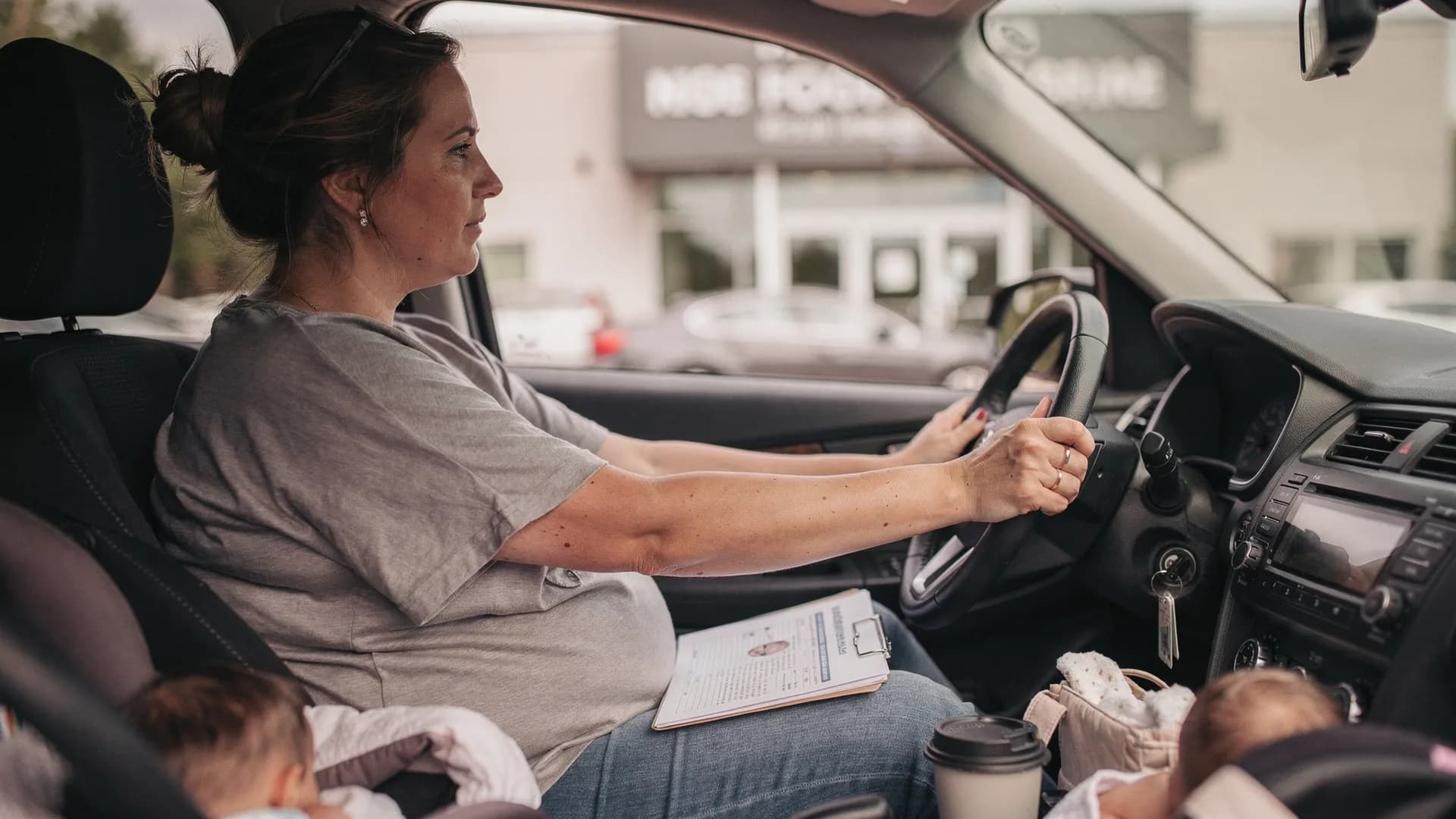 A woman sits in a parked car outside a clinic with her hands resting on the steering wheel.