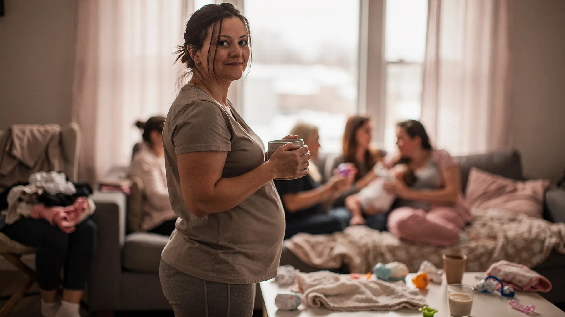 A mother stands apart with a mug while visitors gather around a baby in the background.