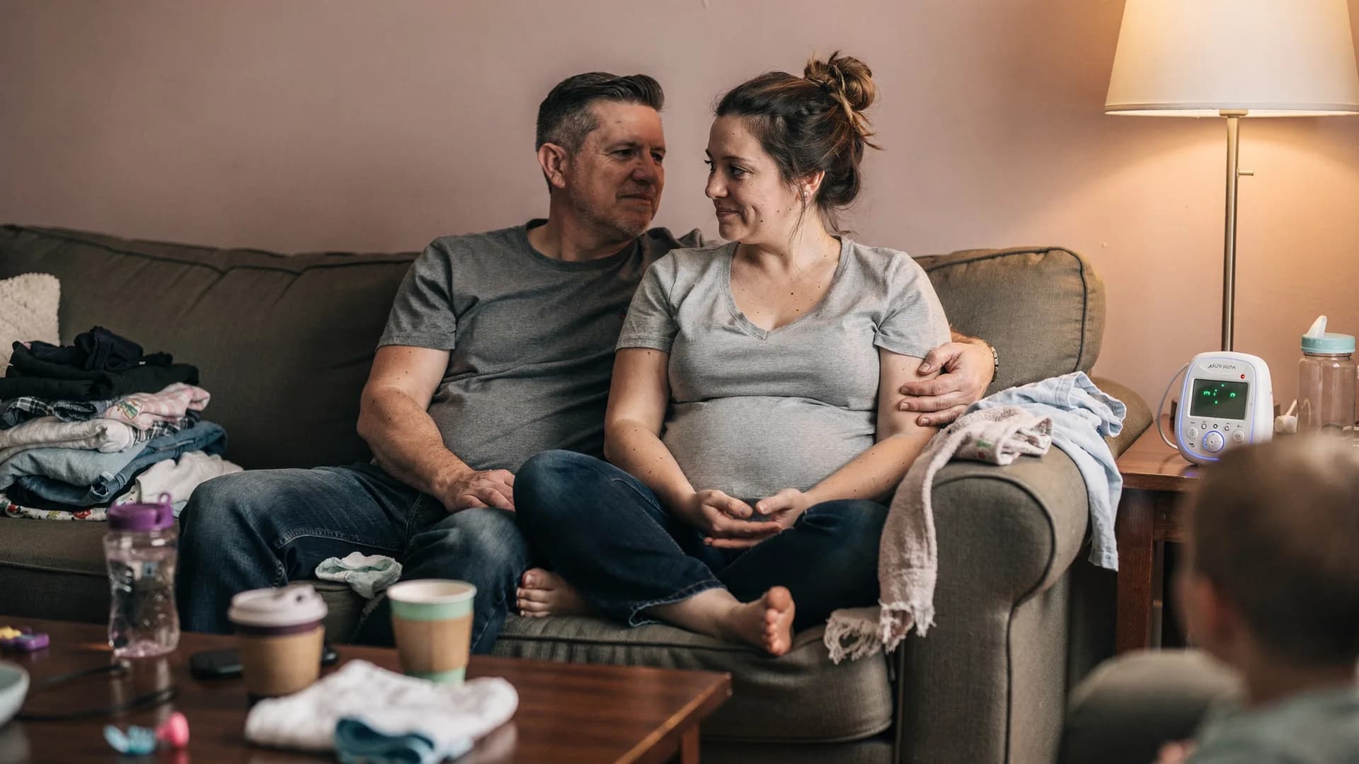 A woman and her partner sit on opposite ends of a couch in warm lamplight.
