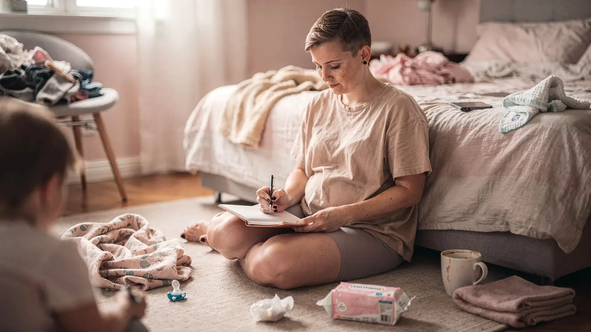 A white postpartum woman with short pixie cut and petite narrow-shouldered body pauses with a pen over an open journal while sitting on a bedroom floor.