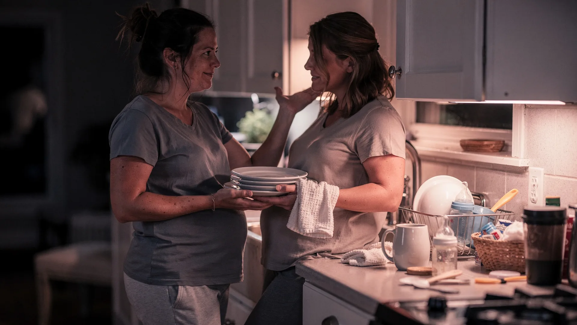 A couple talks quietly in a kitchen after bedtime with dishes and bottles nearby.