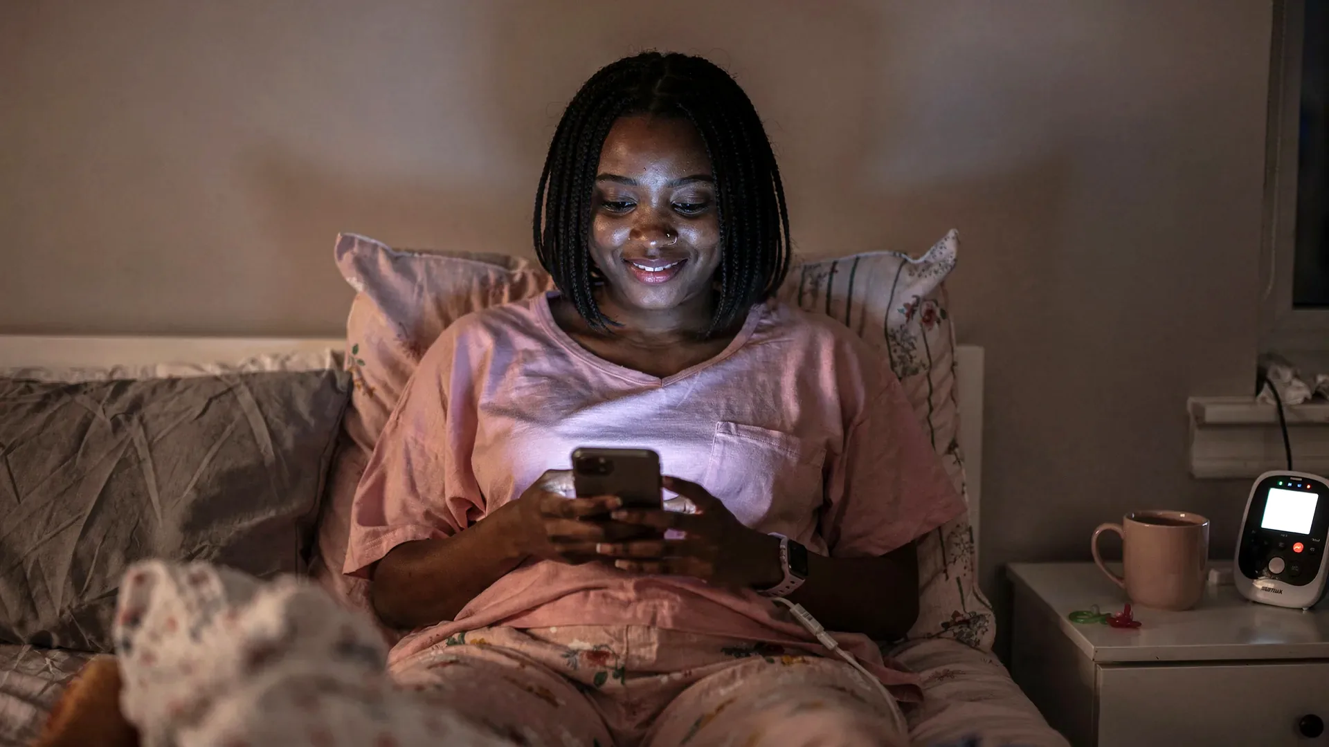 A tired Black postpartum woman with braided bob and petite body smiles at her phone in bed at night with a baby monitor nearby.