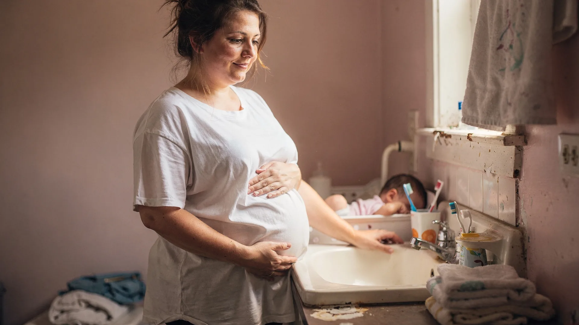 A postpartum woman in an oversized t-shirt stands by a bathroom mirror with one hand resting on her belly.