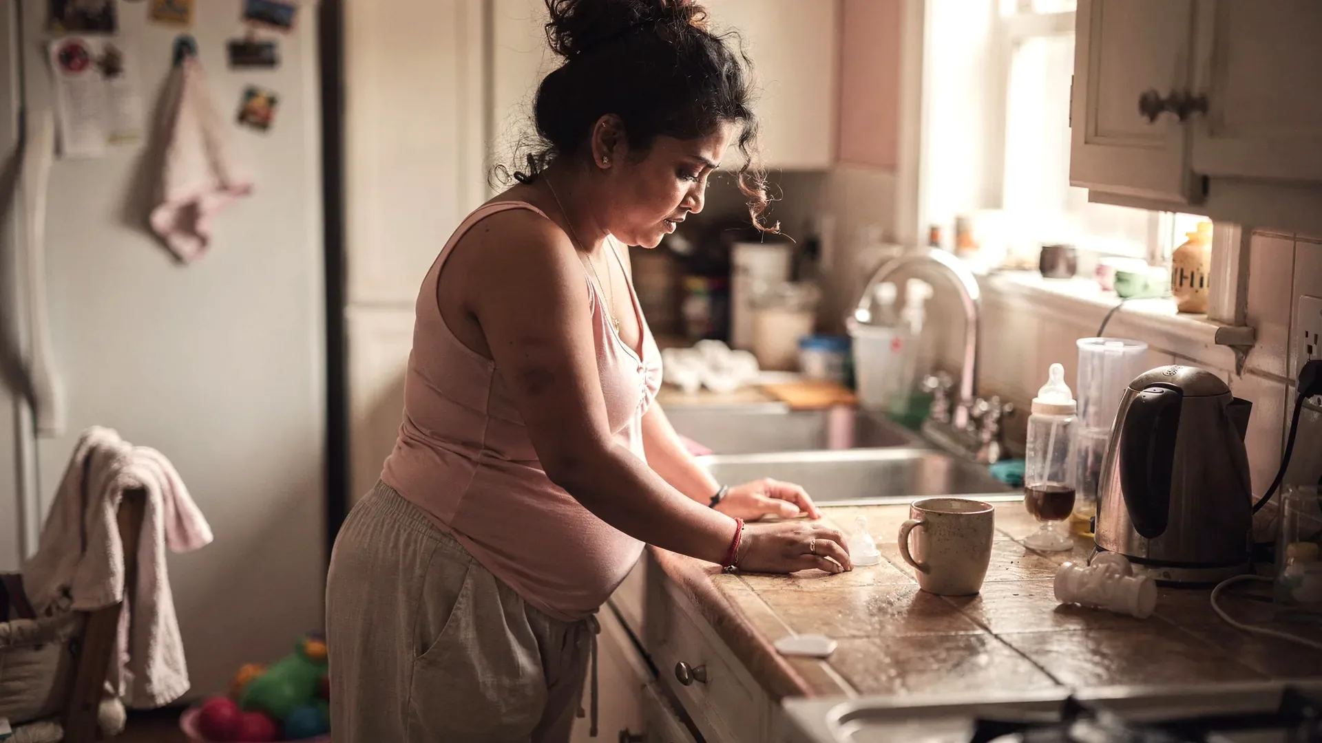 A South Asian postpartum woman with curly dark hair and average body grips a kitchen counter beside scattered toys and a kettle after a hard moment.