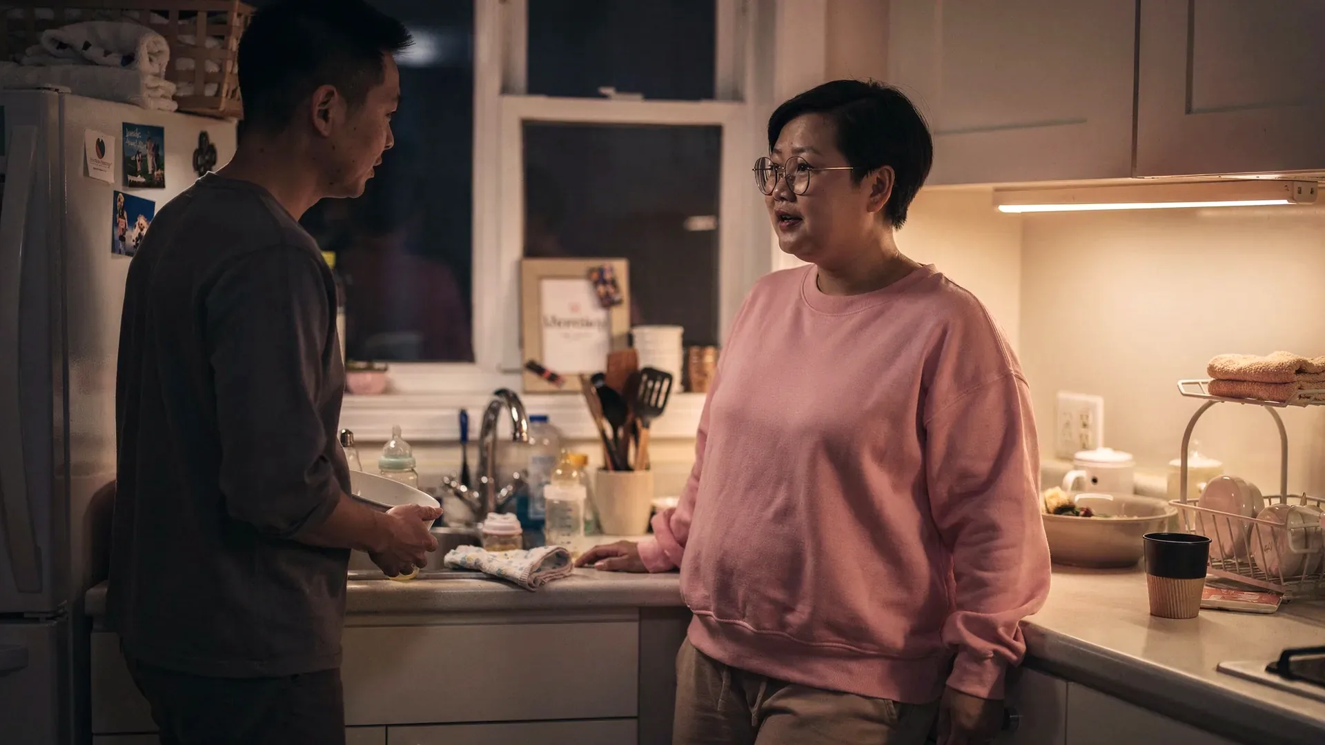 An East Asian postpartum woman with pixie cut and curvy postpartum body and her partner talks quietly in a kitchen after bedtime with dishes and bottles nearby.