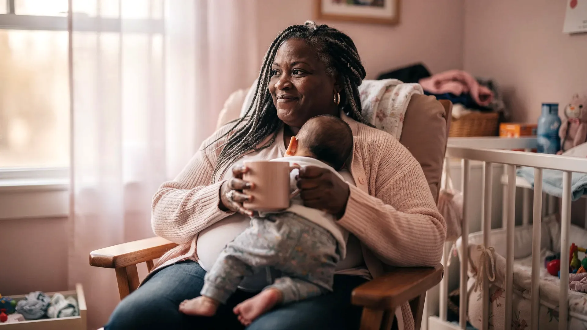 A Black postpartum woman with gray-streaked braids and curvy body sits in a nursery chair holding a mug while a baby rests turned away on her shoulder.